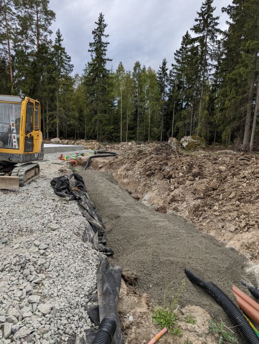 Grundarbete för husbygge med rörgrav, grus och makadam, samt en grävmaskin i bakgrunden.