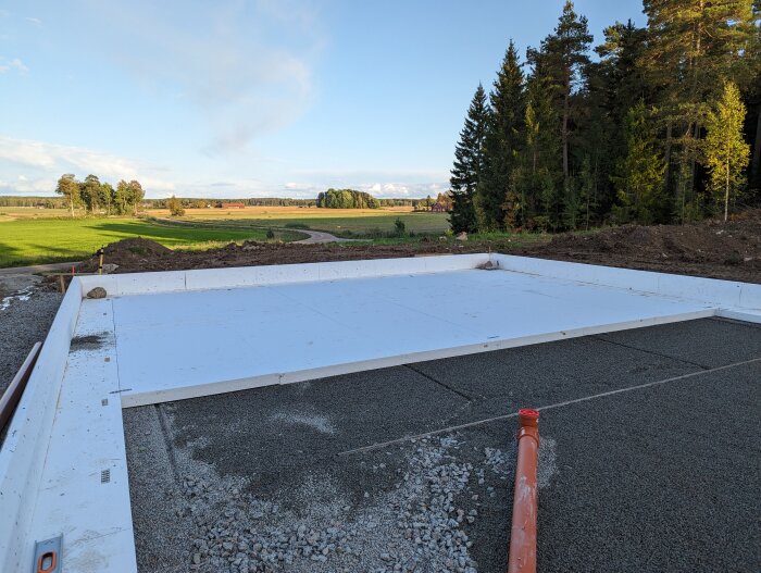 Grund med kantelement och synliga avloppsrör på byggarbetsplats omgiven av landskap och träd.