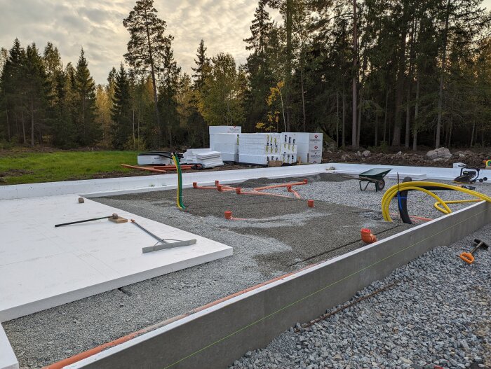 Grundarbete för bostadshus med kantelement, avloppsrör, makadam och verktyg framför skogsbakgrund.