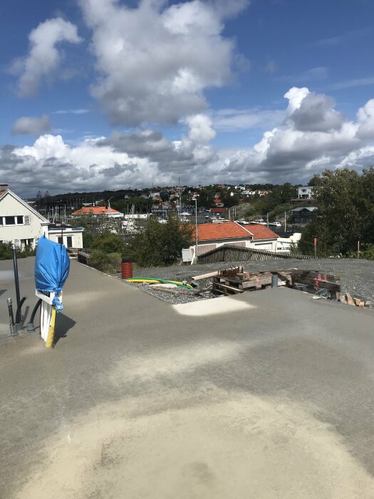 Utsikt över ett byggarbetsområde med material och utrustning, hus och segelbåtar i bakgrunden.