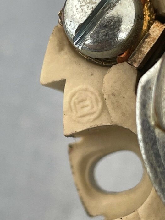 Närbild på en symbol som ser ut som ett "B" ingraverat i en beige yta, intill en skruv och en speglande yta.