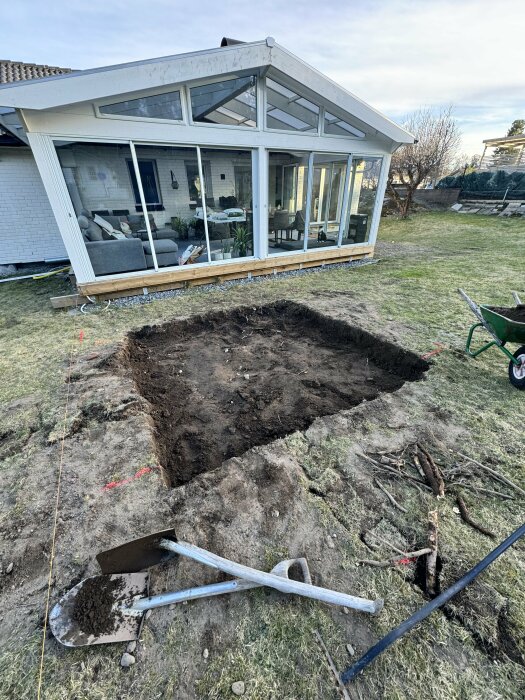 Uppgrävd jord framför en glasveranda, borttagna rötter syns med spade och skottkärra.