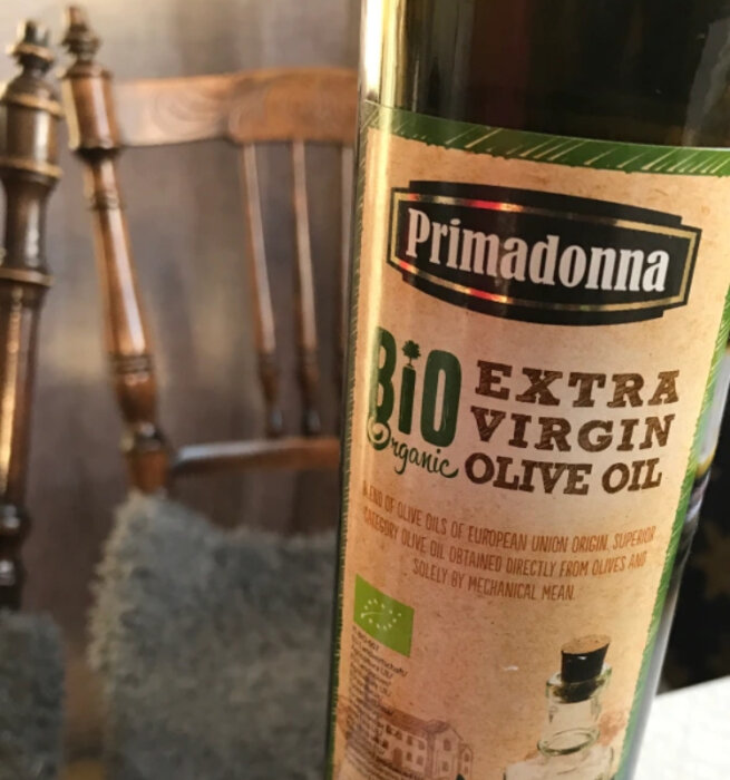 Flaska med 'Primadonna Bio Extra Virgin Olive Oil' framför en suddig stol och matta.