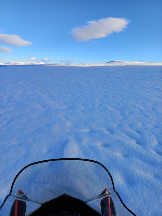 Vy från en snöskoter som färdas över ett snötäckt landskap med fjäll i fjärran under en klarblå himmel.