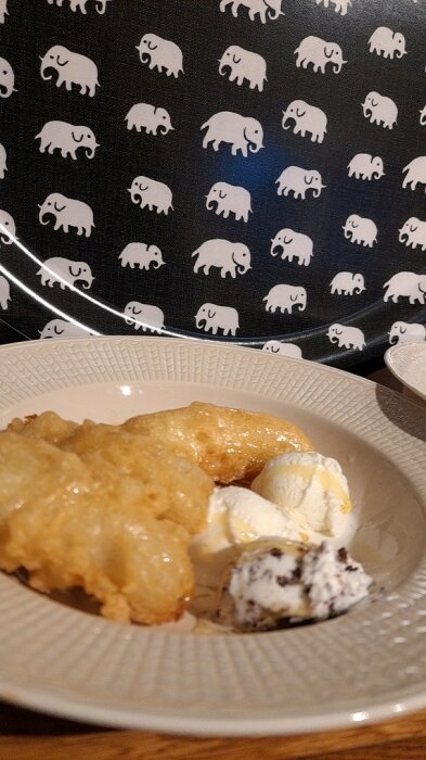 Friterad banan med vaniljglass och sirap på en tallrik, bakgrund med elefantmönster.