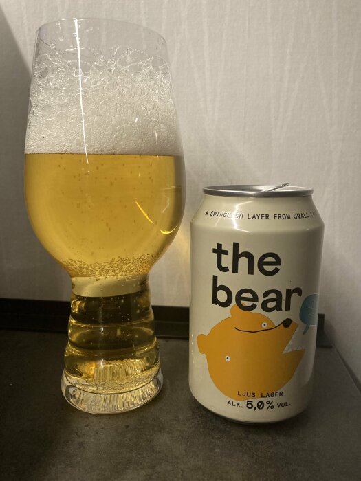 Ölglas fyllt med gul öl bredvid en burk med texten "the bear", ljus lager 5,0% volym med björnmotiv.