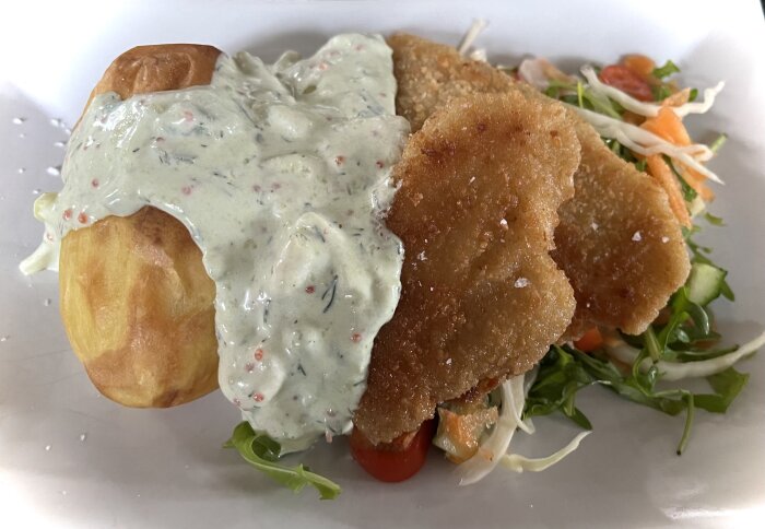 Panerad plattfisk med bakad potatis täckt av räk- och dillsås, serverad med sallad, avokado och lök.