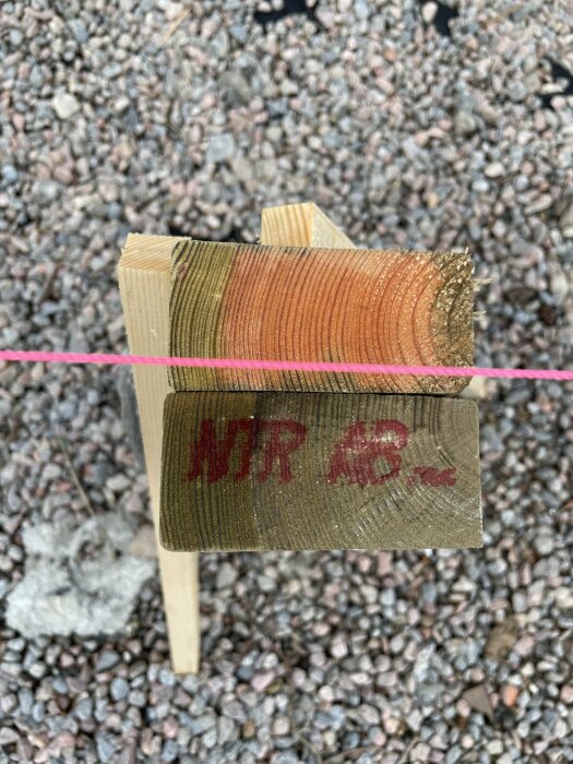 Träbit med stämpeln "NTR AB" mot en bakgrund av grus och ett rosa snöre.