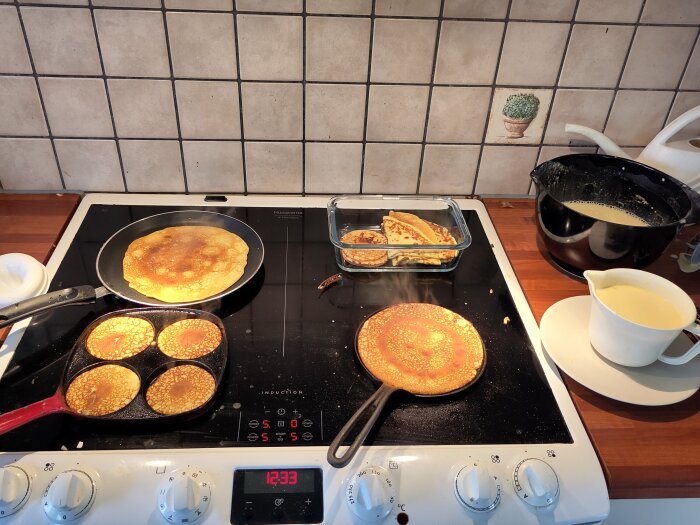 Stekning av både stora och små pannkakor på en spishäll, med färdiga pannkakor i en ugnsform.