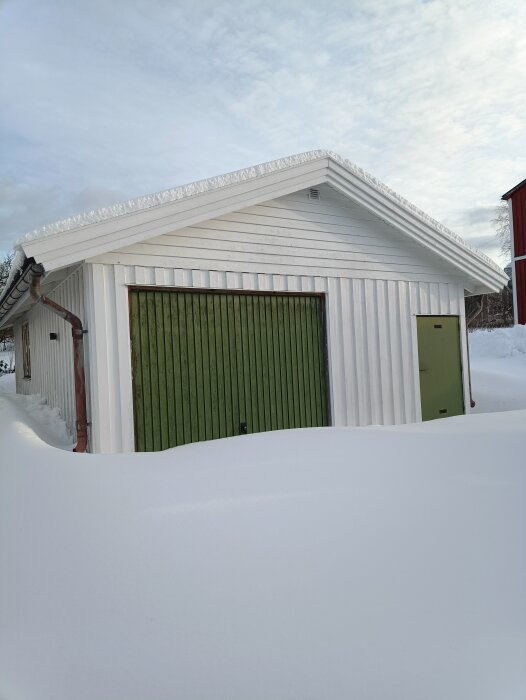 Ett vitmålat garage med nytt plåttak, grön port och omgivet av snö under en himmel med lätt molnighet.
