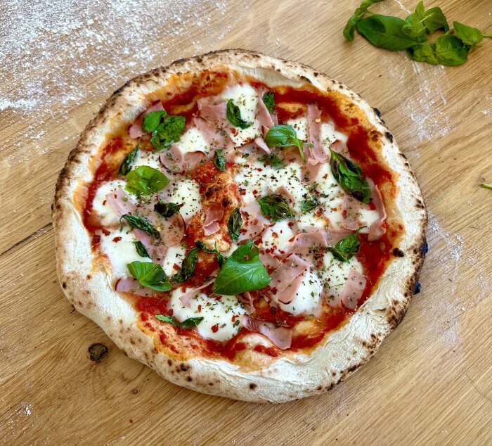 Hemgjord pizza med tomatsås, ost, skinka och basilika på träbord med mjölströdd yta.