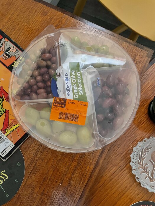 Förpackning med sorterade gröna och lila oliver på bord, 30% rabattetikett synlig.