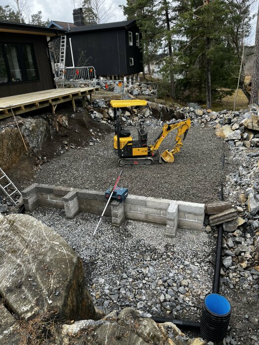 Formbygge av grå betongblock för platta nära en husgrund med grävmaskin och byggmaterial.