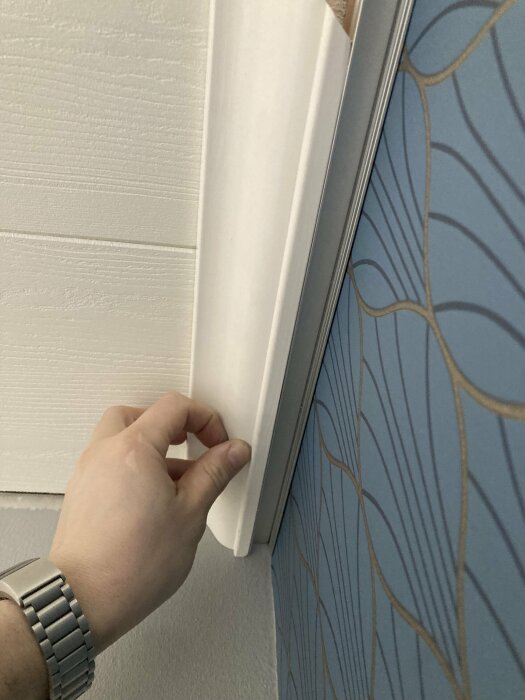 Hand som justerar vita lister mot en vägg med blått tapetmönster.