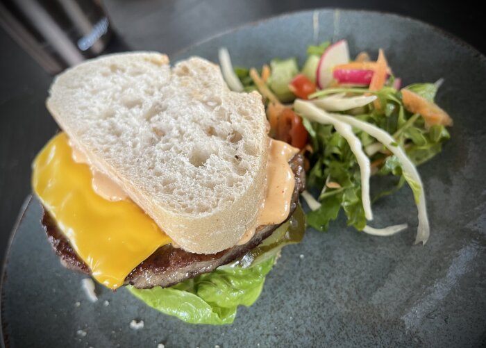 Hamburgare med ost och dressing mellan ciabattabröd, serverad med sidosallad på en grå tallrik.