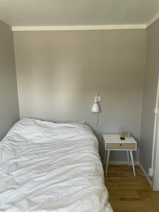 En hörna av ett sovrum med en säng och ett litet nattduksbord med en lampa, visande golv och väggmatta.