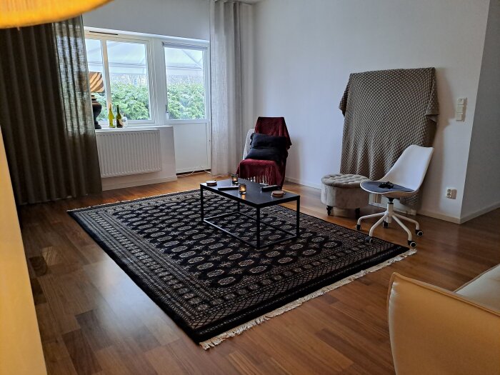 Vardagsrum med matta, en fåtölj, ottoman, vit stol, fönster och en draperad filt över högtalare.