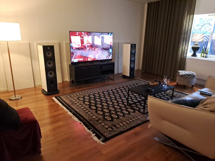 Vardagsrum med två golvstående högtalare, TV, soffa, svart bord på persisk matta, stående lampa och fönster med gardiner.