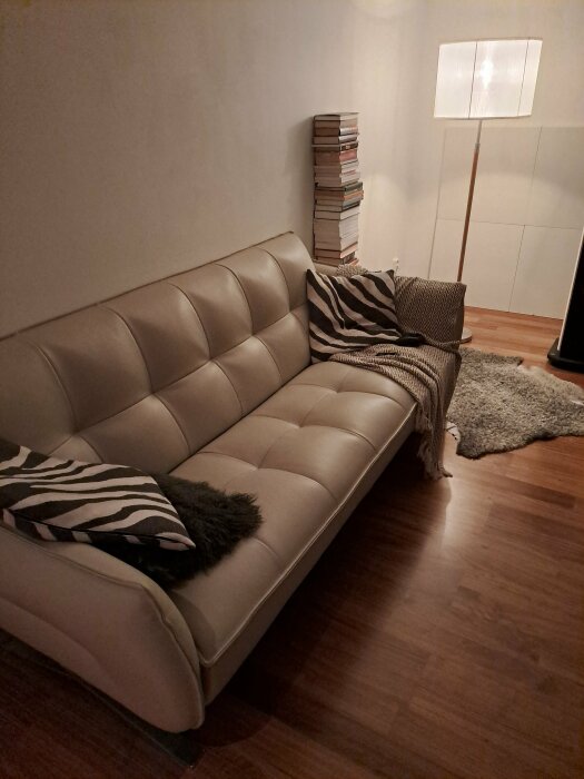 Minimalistiskt vardagsrum med en beige soffa, dekorativa kuddar, en stående lampa och en stapel böcker.