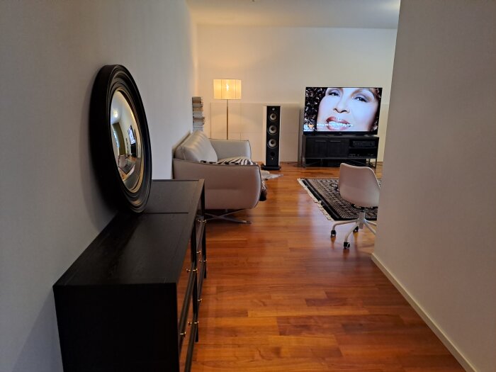 Ett modernt vardagsrum med en beige soffa, två vita stolar, tv på väggen, trägolv, svart möbel och en rund spegel.
