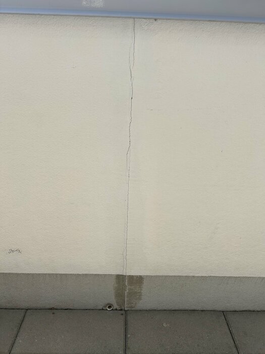 Vertikal spricka längs en vägghörn ovanför ett grått kaklat golv på en uteplats.