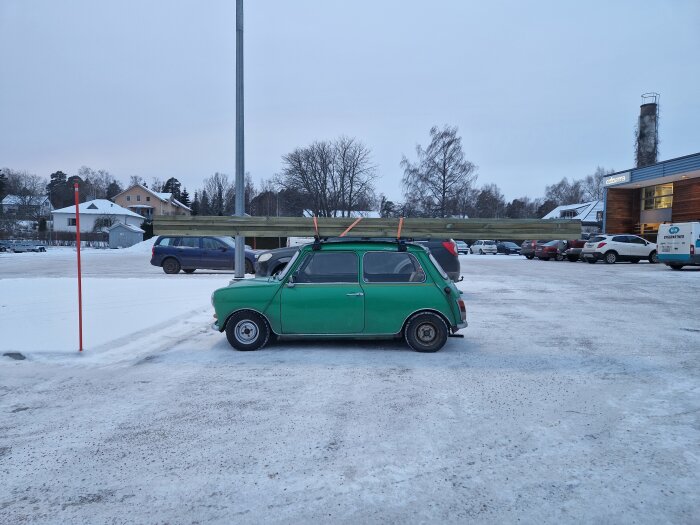 Liten grön bil på parkering med lång impregnerad bräda ovanpå i vinterlandskap.