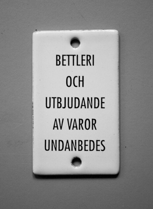 Emaljskylt med texten "BETTLERI OCH UTBJUDANDE AV VAROR UNDANBEDES" på en grå bakgrund.