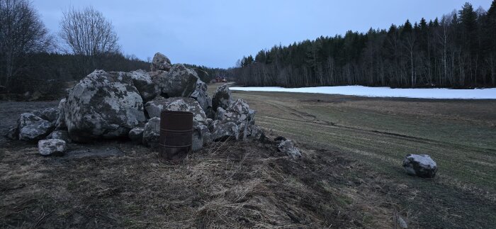 Hög med stora stenar på ett fält med synliga hjulspår och kvarvarande snö i bakgrunden.