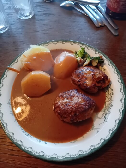 Tallrik med två köttbullar, potatis och brunsås, bestick i bakgrunden.