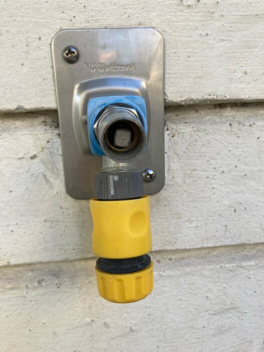 Vattenkran på husvägg med påkopplad slangkoppling av gul och svart plast.