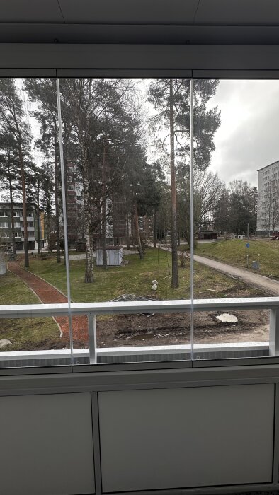 Vy genom fönster över en park med träd och promenadvägar mellan bostadshus.