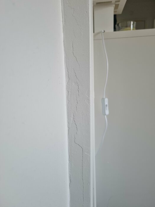 Spricka i en vitmålad vägg vid dörrkarm med spackel synligt och en kabel längs med karmen.