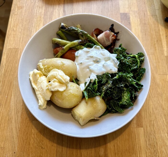 En tallrik med grillunch bestående av kokt potatis, grillad sparris och salladskål med vit sås.