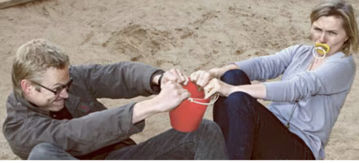 Två personer drar i en röd hink på en sandstrand, kvinnan har en spade i munnen.