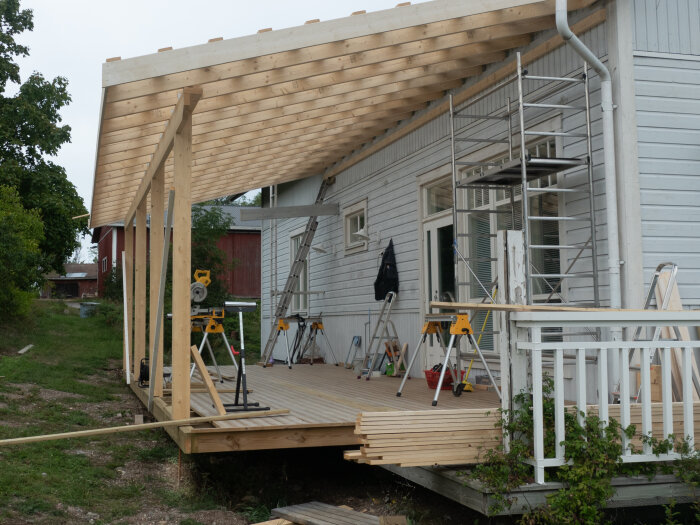 Nybyggd träaltan vid hus med stöd av gängad stålstång, byggmaterial och verktyg synliga.
