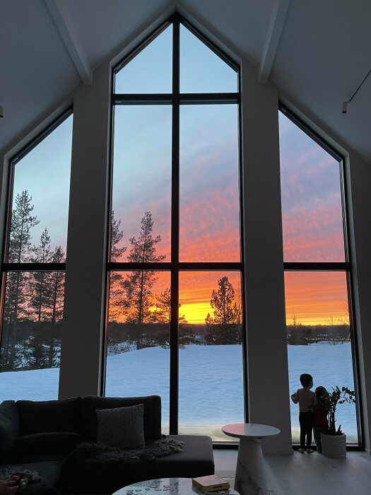 Inomhusvy med stora fönsterpartier som visar en solnedgång med orange himmel och snötäckt landskap, två barn framför fönstret.