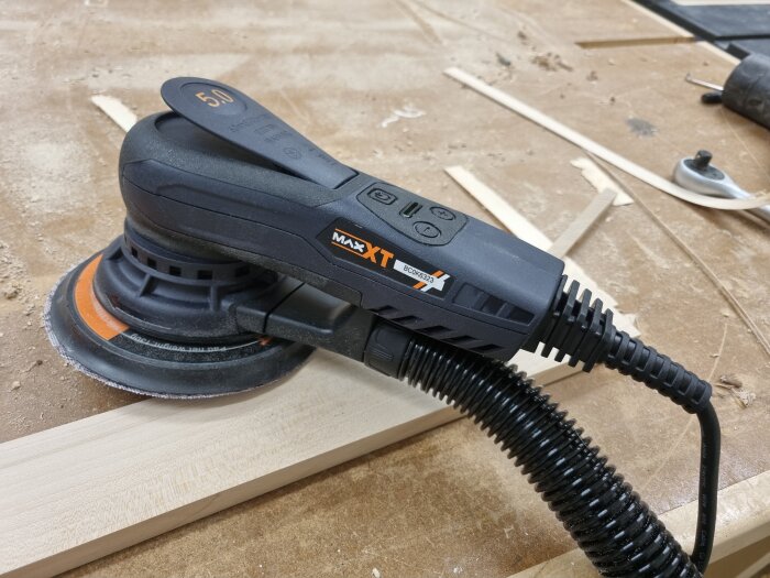 Slipmaskin på ett träarbetsbord med ansluten dammutsugsslang, omgiven av träspån och verktyg.