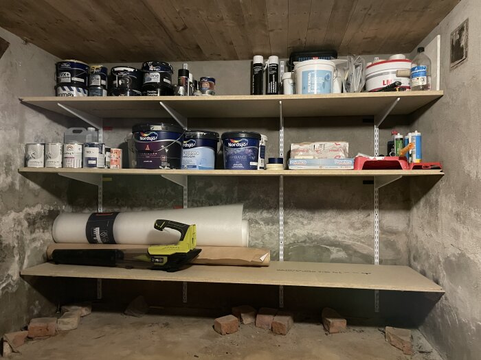 Hyllor i källare fyllda med färgburkar, byggmaterial och verktyg.