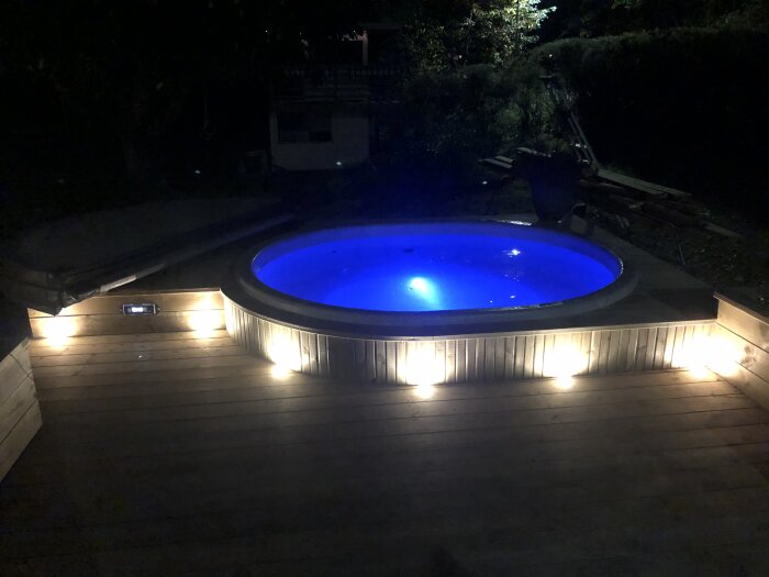 Upplyst rund pool i mörker med träaltan och integrerad belysning.