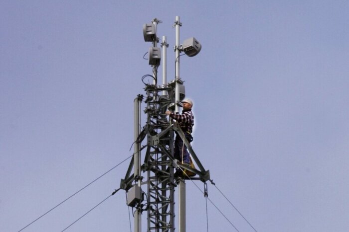 Arbetare klättrar på mobilmast fylld med antenner mot blå himmel.