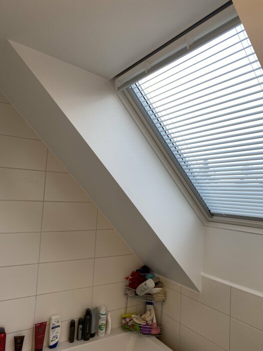 Nymålat snedtak och fönster med persienner i ett badrum, och hyllor med badrumsprodukter.