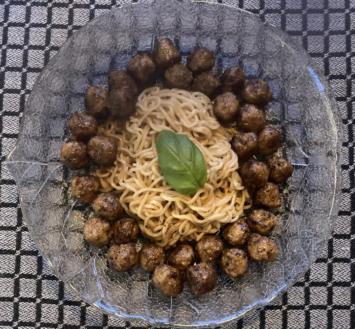 Tallrik med köttbullar och nudlar på rutig duk, toppat med ett basilikablad.