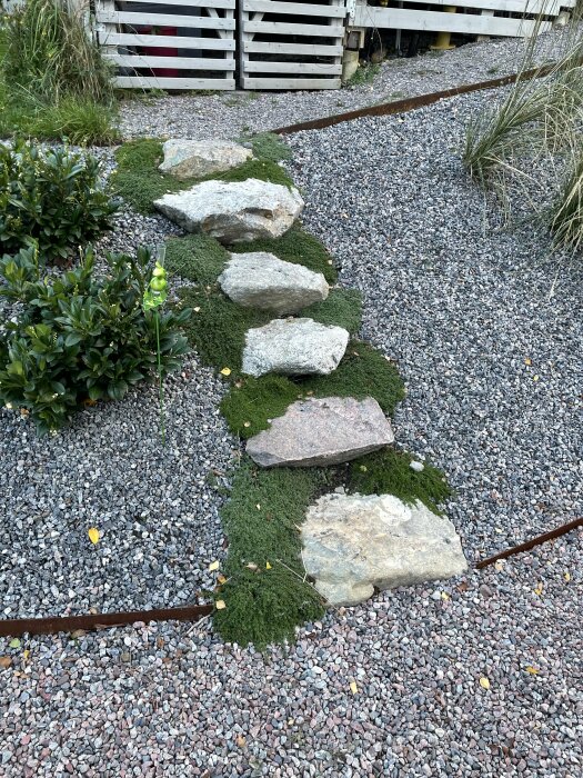 Stensteg snirklar sig genom en slänt med markduk och grå makadam, avdelat med cortenstål, omgivet av gröna växter.
