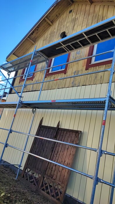 Ställning framför hus med pågående byte av träpanel, gamla paneler lagda vid husets grund.