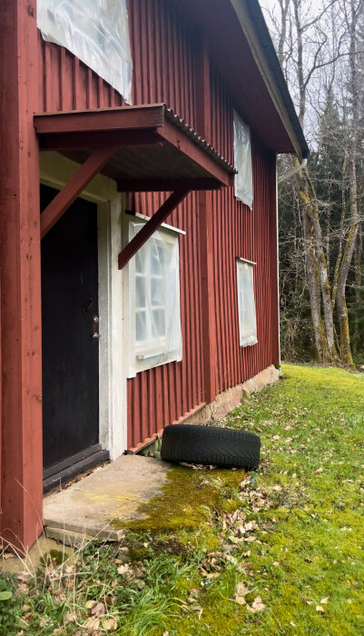 Rödfärgat torp med skadad grund och plasttäckt fönster, omgivet av grönt gräs och skog.