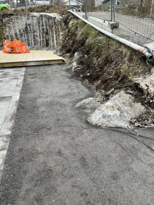 En asfalterad gångväg leder mot en träplattform vid kanten av en slänt med utspridda stenar och en sliten mur i bakgrunden.