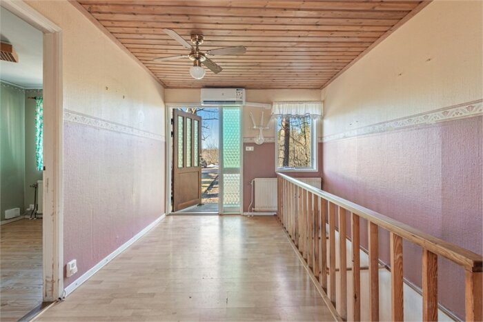 Interiör av en rymlig hall med trägolv, ljusrosa väggar och takfläkt, med utsikt mot dörren.