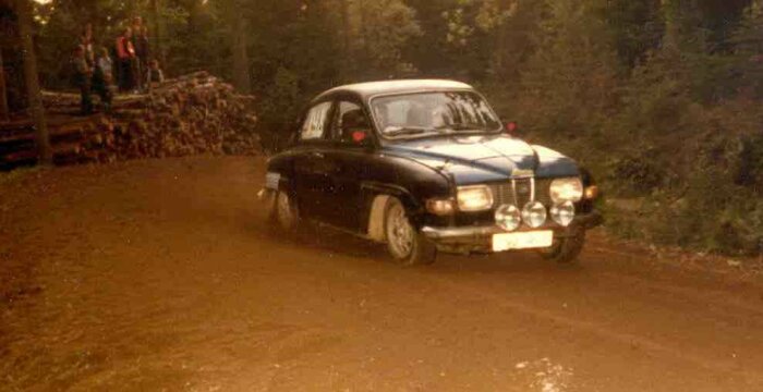 SAAB V4 raliybil som kör på en grusväg med tittare i bakgrunden.