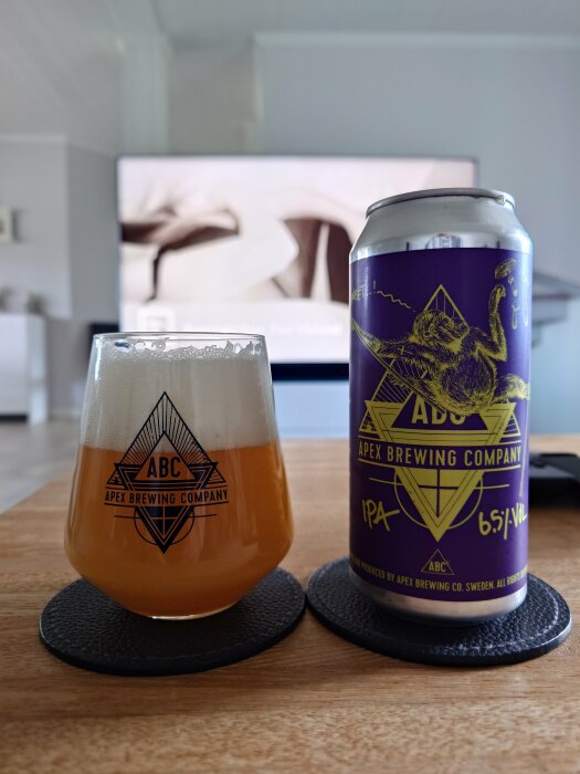Ölglas märkt 'Apex Brewing Company' och en ölburk med vargdesign, på bord framför suddig TV-skärm.