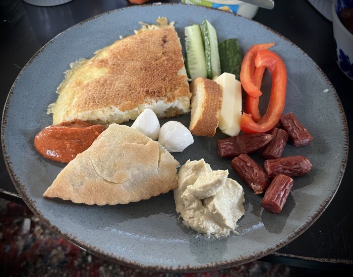 Tallrik med omelett, hummus, skivor av ost och gurka, röd paprika, vitlöksklyftor och kabanos på en bordduk.